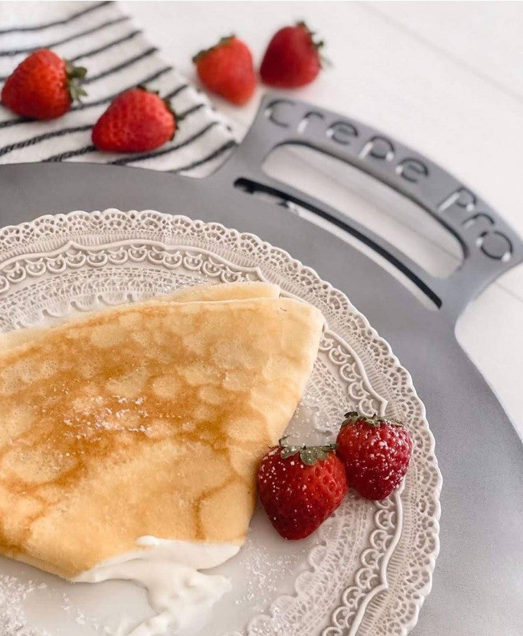 Crepe with whipped cream and strawberries on a decorative plate with a Crepe Pro machine in the background. Carbon steel pan crepe pan cooking crepes on stovetop