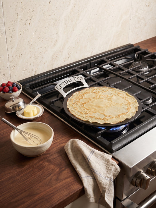 A cast iron skillet with a 'Crockpot Pro' handle sits on a lit gas stove burner, cooking a thin crepe. The scene is set on a dark wood countertop with bowls of batter, butter, fresh berries, and a checkered kitchen towel, against a textured beige tile wall.