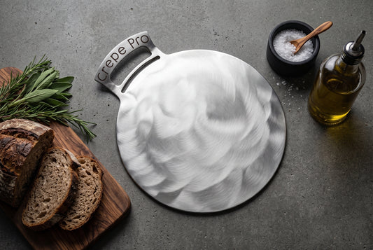 A round hammered stainless steel serving board with a handle engraved with 'Crepe Pro', styled on a gray stone surface with sliced bread on a wooden board, a small bowl of coarse salt, and a bottle of olive oil.