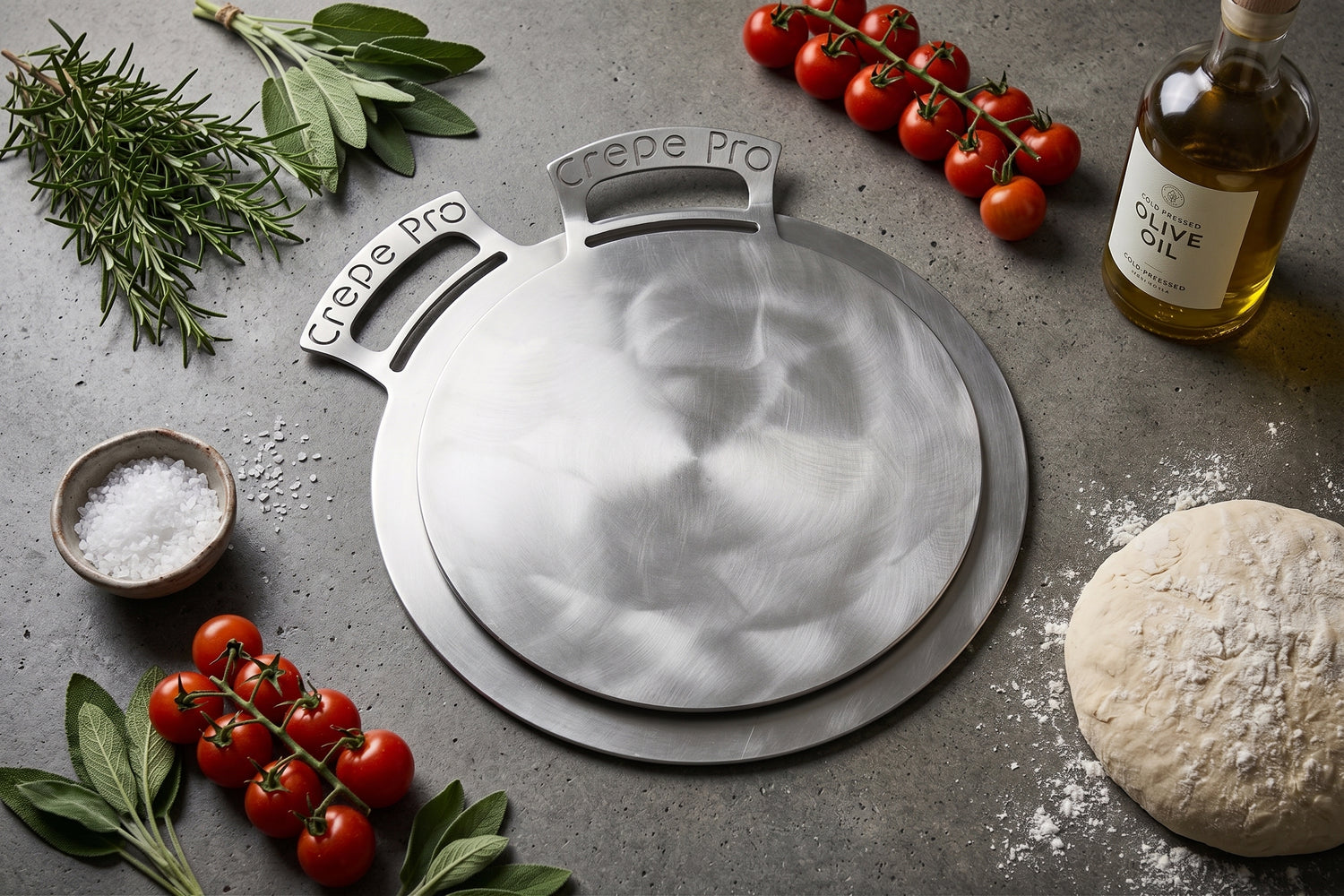 A round stainless steel crepe maker pan with two handles labeled 'Crepe Pro' is shown on a gray stone surface, surrounded by food props including cherry tomatoes, sage, rosemary, olive oil, coarse salt, and dough.