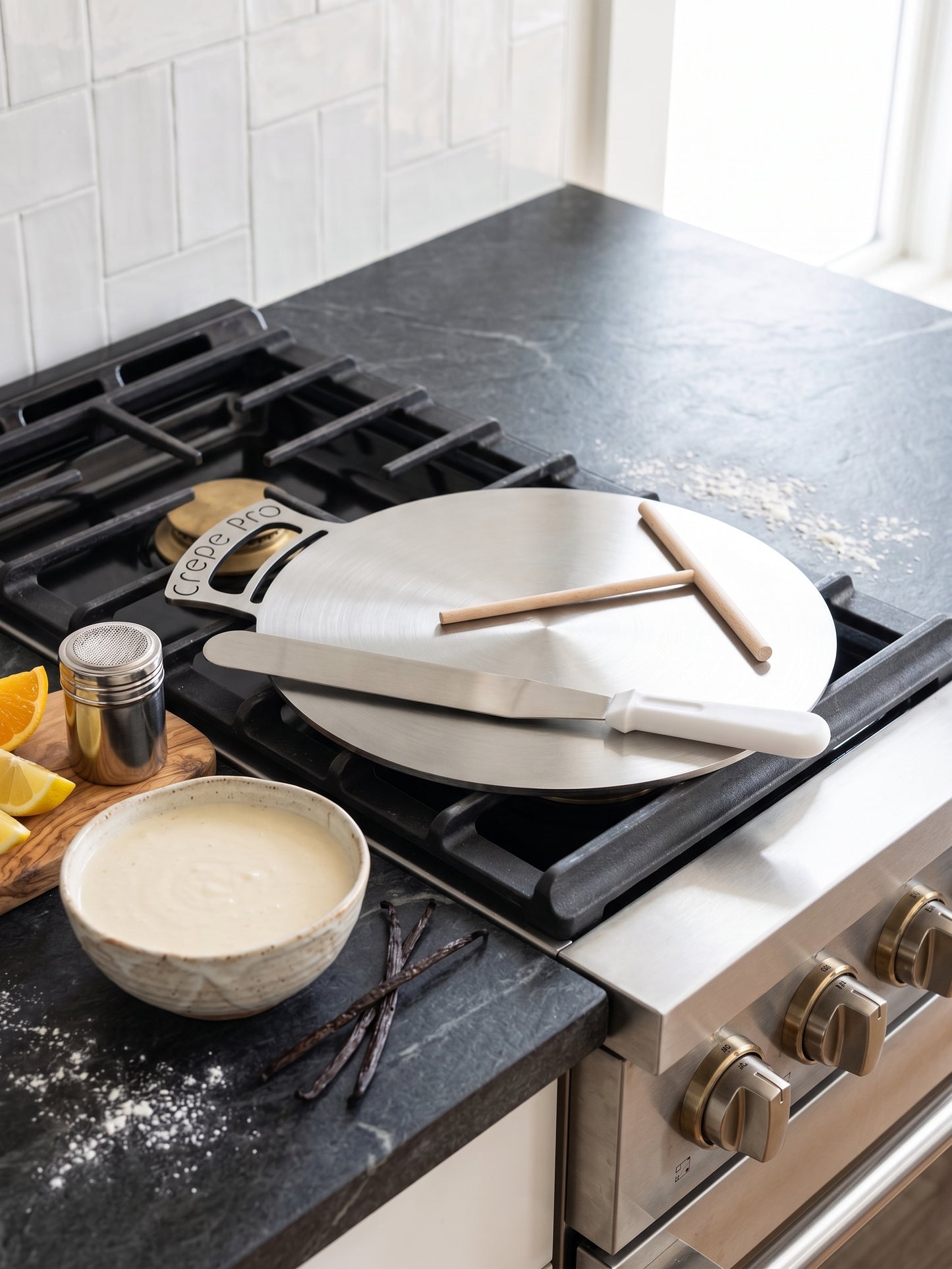 A round metal cake pan with a wooden handle sits on a black gas stovetop in a kitchen. A metal spatula and two wooden sticks rest on the pan, with a small bowl of batter, vanilla beans, and lemon slices nearby on a dark countertop.