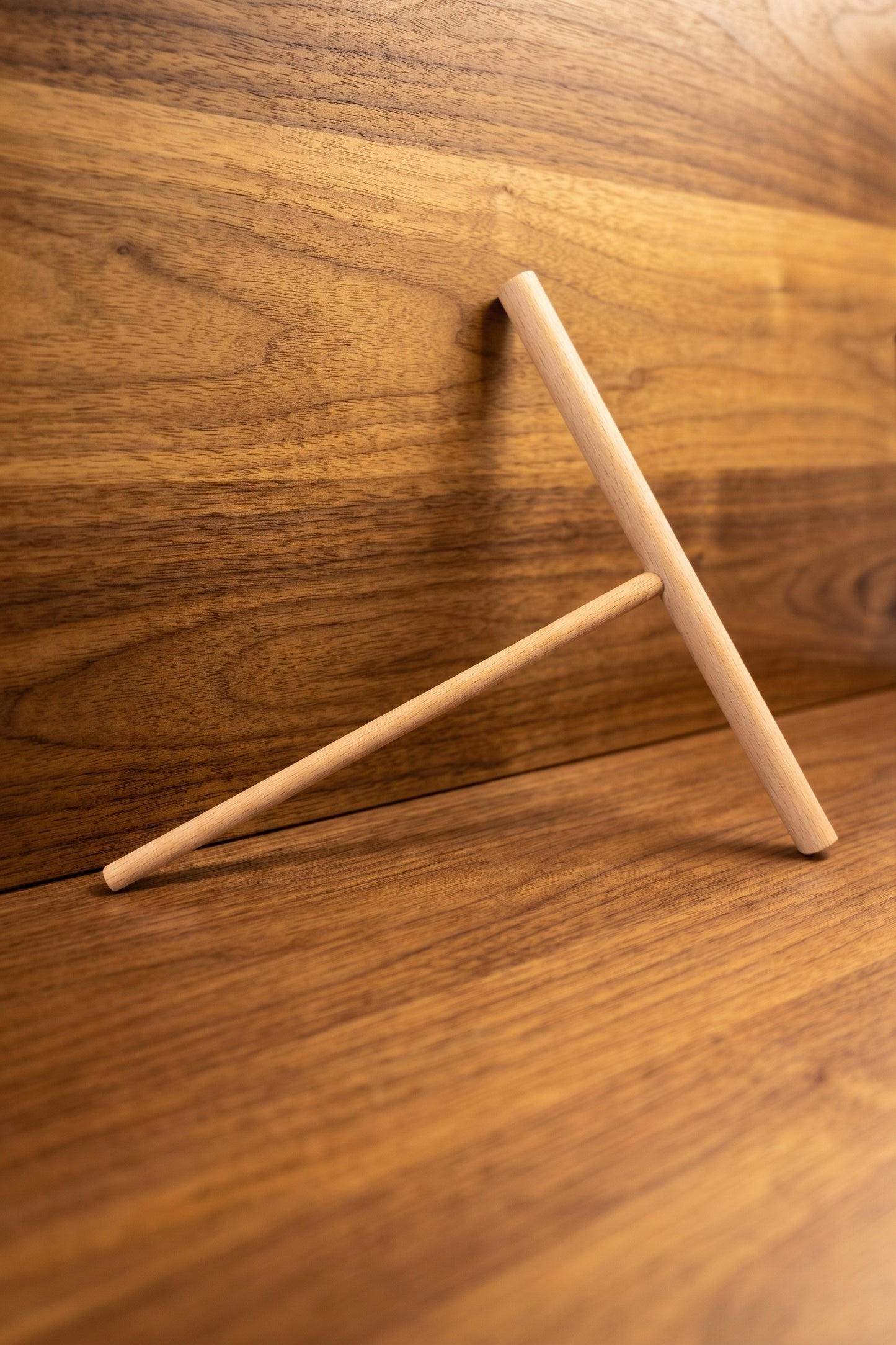 A minimalist wooden T-shaped desk stand made of light natural wood, shown on a matching wood-grain surface with a warm, golden-brown background.